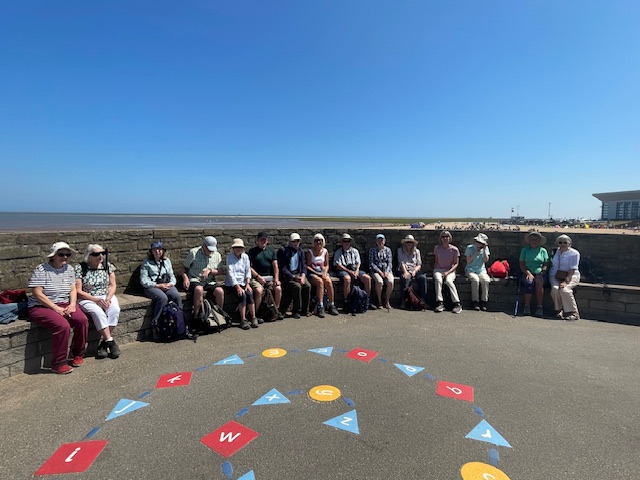 Members sit around a circular stone bench near the beach, many with hats on